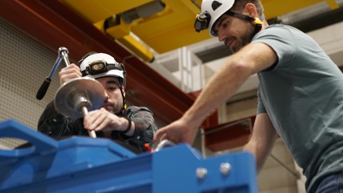 two men winding a metal piece of equipment on a blue frame